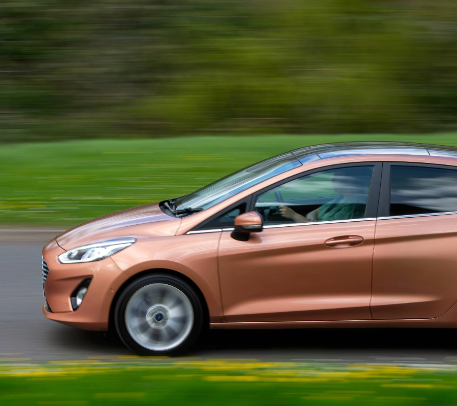 Copper-colored compact car driving at speed on a road with green grass in the background