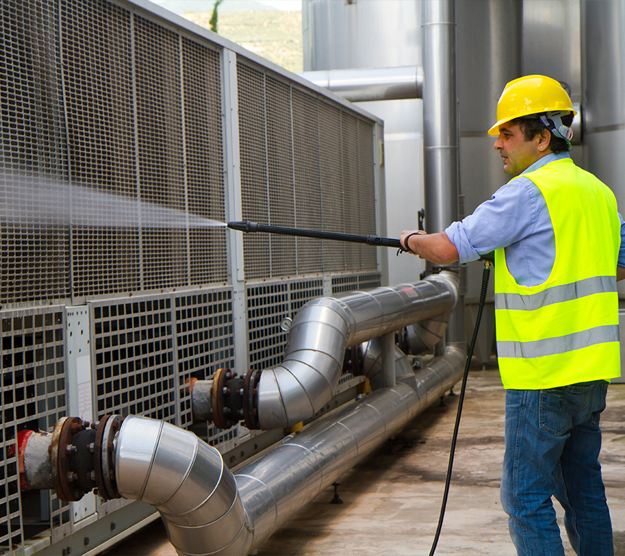 A man in a yellow vest cleans a large industrial air conditioner, ensuring it runs efficiently and stays in top shape.