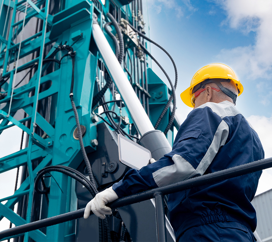 A full color image of a man wearing a hard hat and safety glasses stands beside a drilling rig.