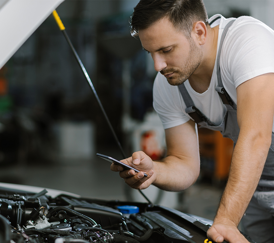 A man checks his phone as he repairs a car, leveraging digital tools for hands-on industrial work.