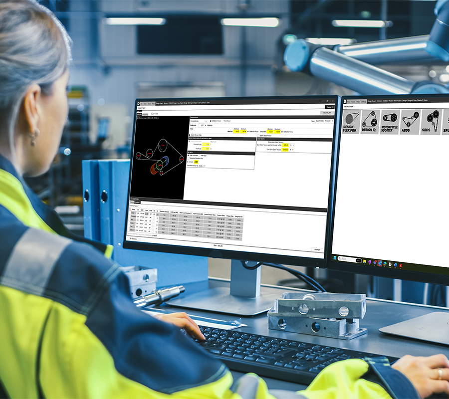 A woman in a safety vest analyzes power transmission drive data on two computer screens in an industrial setting.