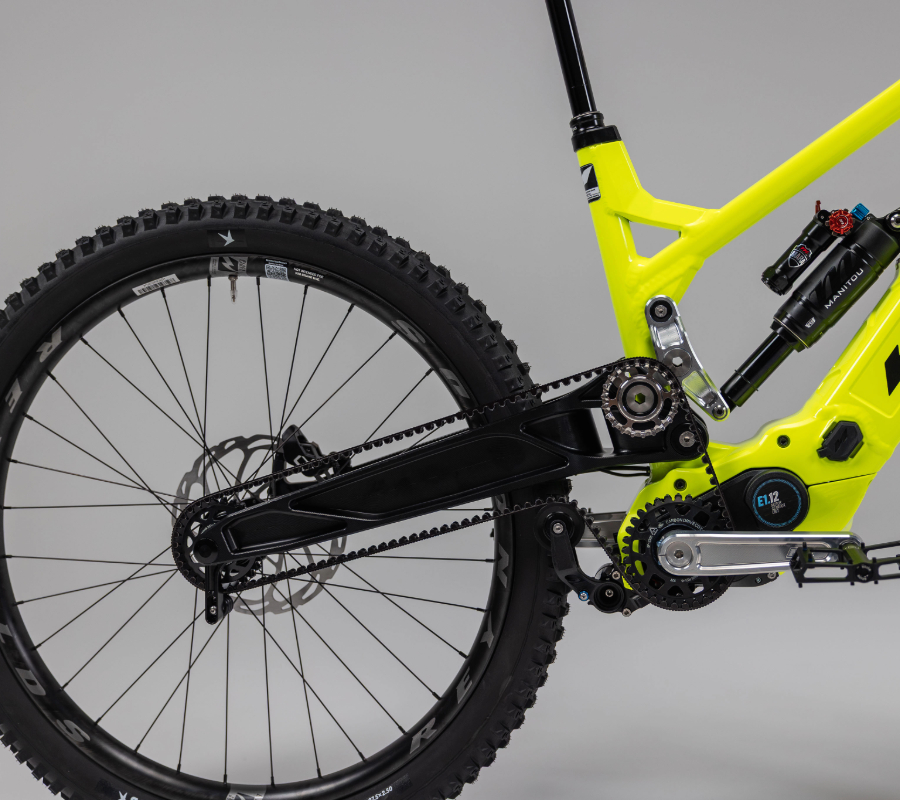 Rear section of a neon yellow full-suspension e-mountain bike showing the E1.12 mid-drive motor, belt drive, rear disc brake, and knobby tire.