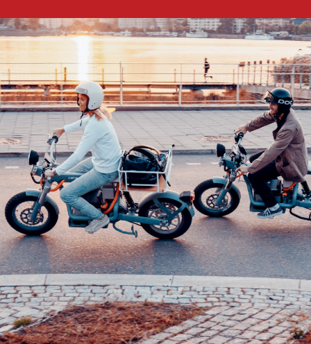 two people ride electric motorcycles along a waterfront street during sunset