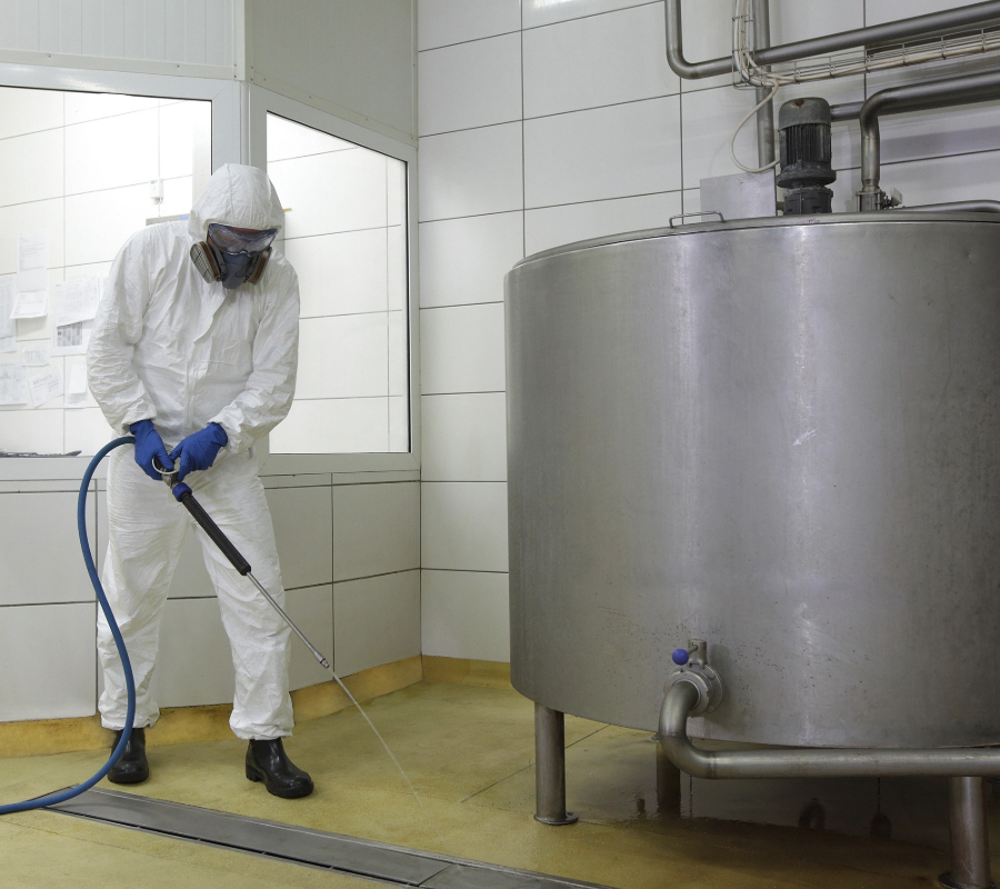 Worker in protective gear using industrial hose for cleaning inside a manufacturing plant.
