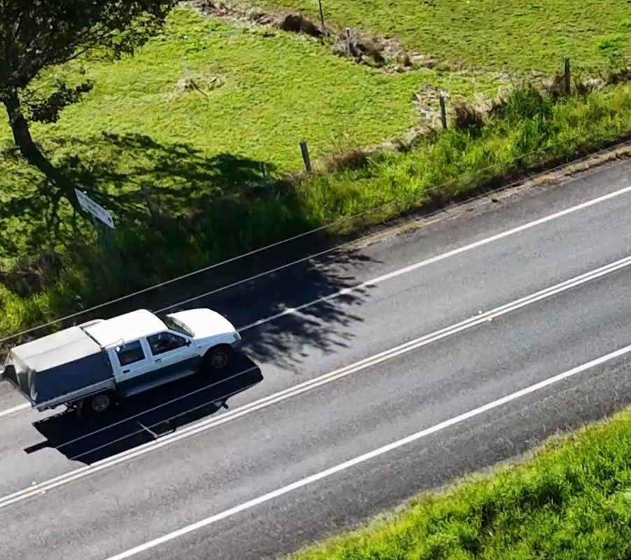 White utility pickup truck driving along a rural road bordered by green fields and trees.