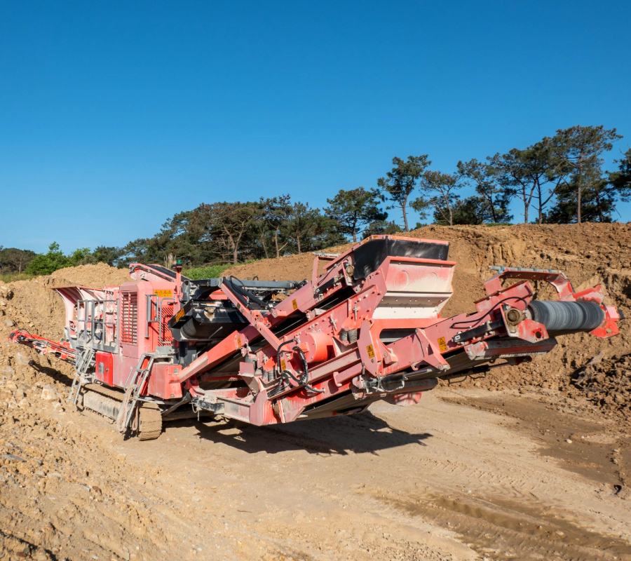 Large red mining machine with conveyor belts operating on a dirt site under clear blue sky.