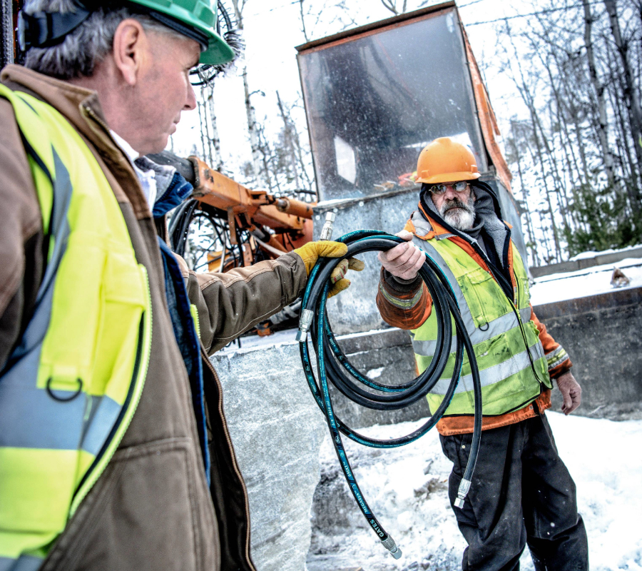 Two construction workers in safety gear exchanging a hydraulic hose at a snowy job site with heavy machinery in the background