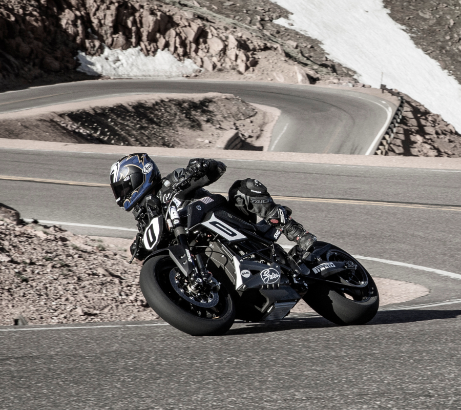 A motorcyclist leans into a sharp turn on a winding mountain road, riding on an electric motorcycle