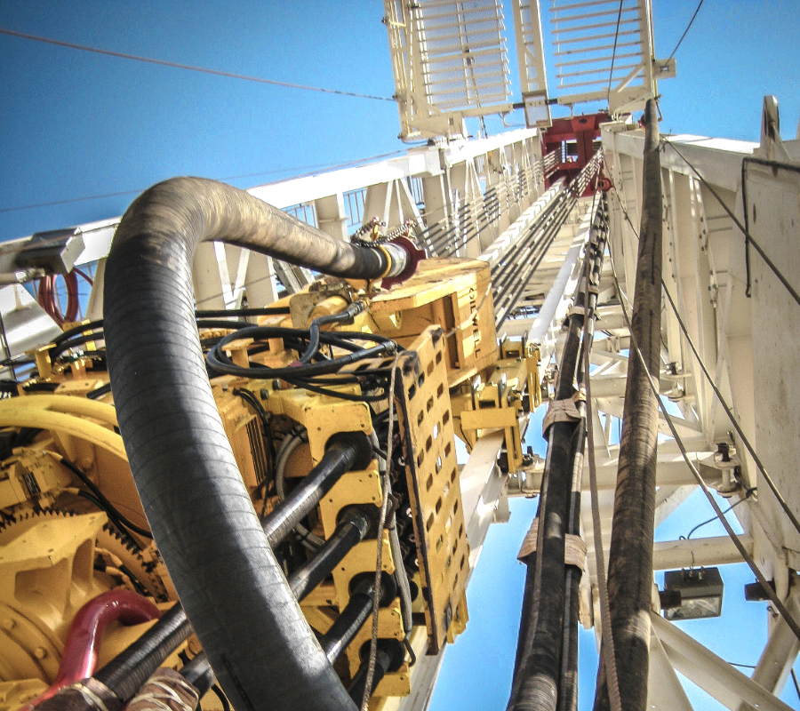 Low-angle view of drilling rig machinery with thick black hoses and yellow components extending upward along the tower.