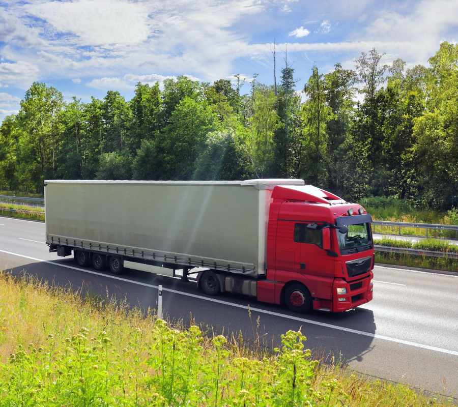 Red semi-truck with a trailer driving on a highway next to green trees.