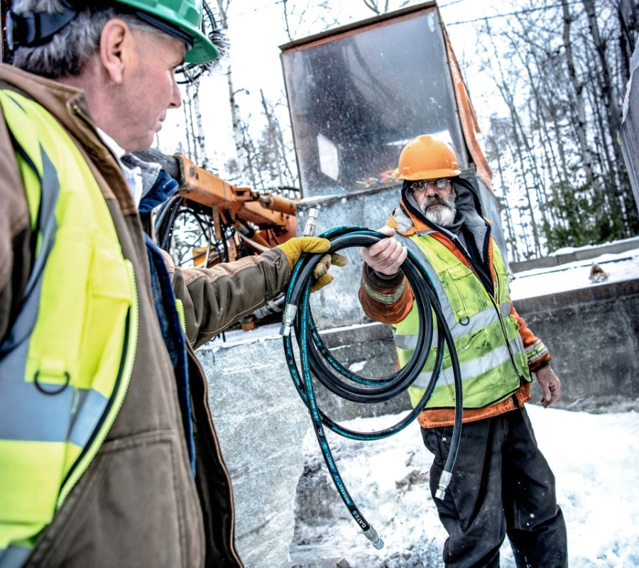 Construction worker in safety gear holding a coiled Gates MXT hydraulic hose on a snowy jobsite.